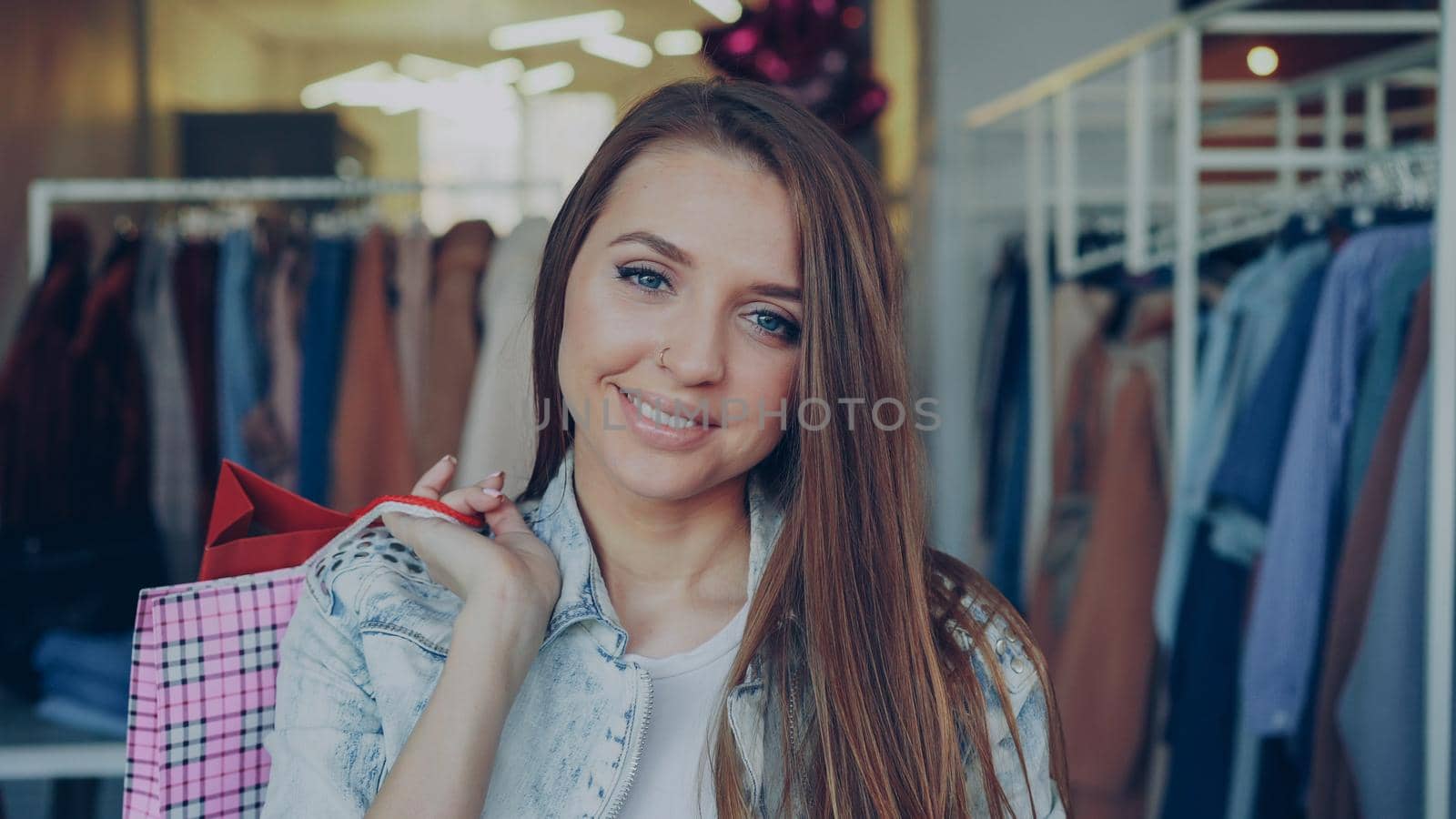 Close-up portrait of attractive young girl standing with paper bags in clothing shop and looking at camera smiling. Stylish women's clothes is in background. by silverkblack