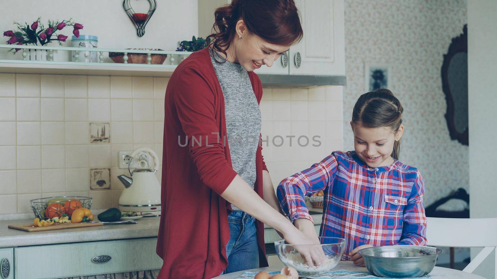 Little cheerful girl helping her mom in the kitchen mixing dough for cookies. Family, food, home and people concept by silverkblack