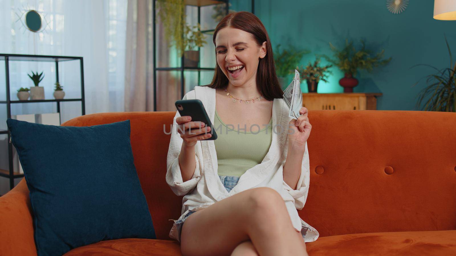 Planning family budget. Smiling lovely girl counting money dollar cash, use smartphone calculate domestic bills at home. Joyful young woman satisfied of income saves money for planned vacation, gifts