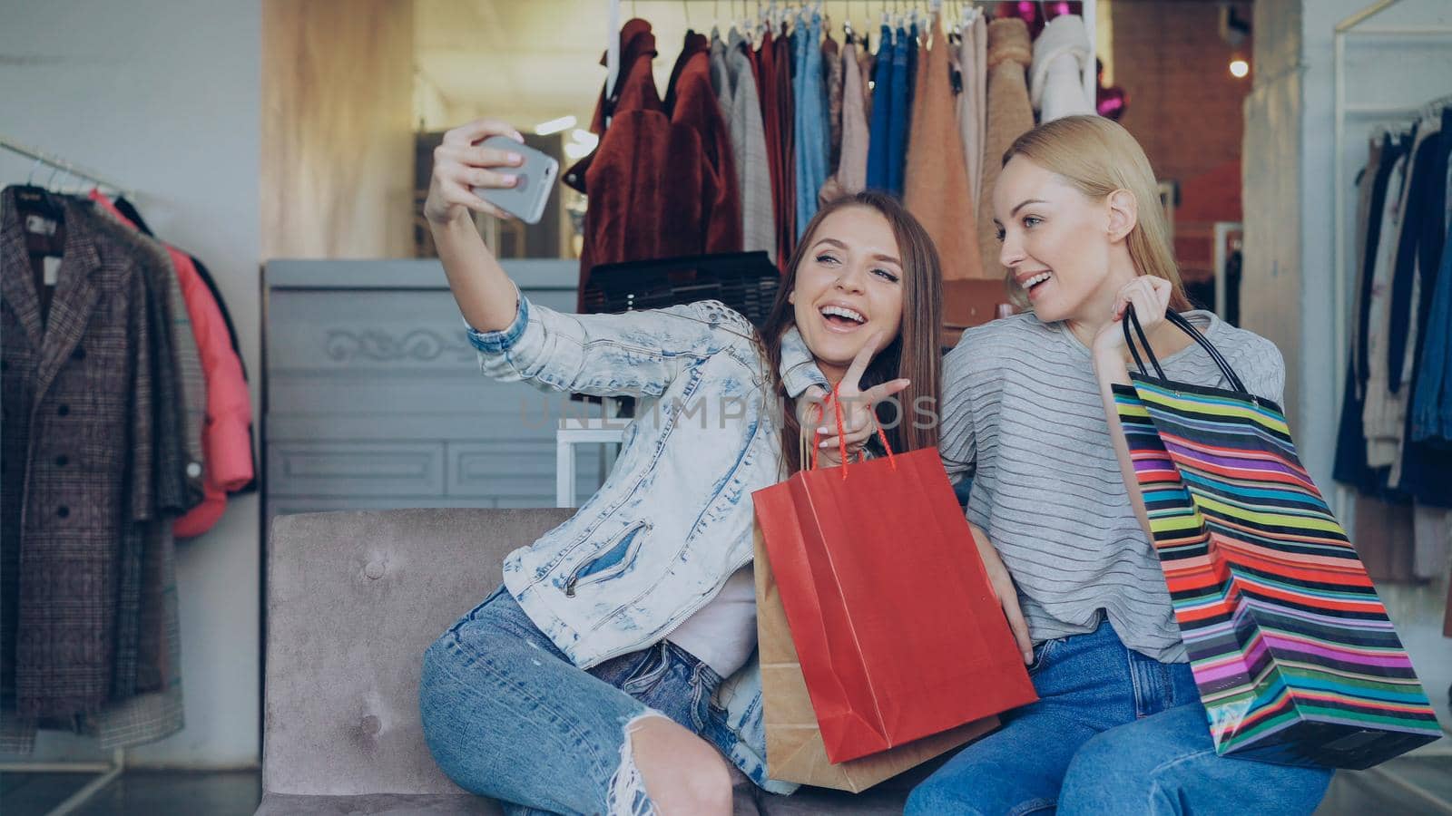 Cute girls are making selfie with colourful paper bags in women's clothing boutique using smartphone, then watching pictures together. They are smiling and laughing caresessly. by silverkblack