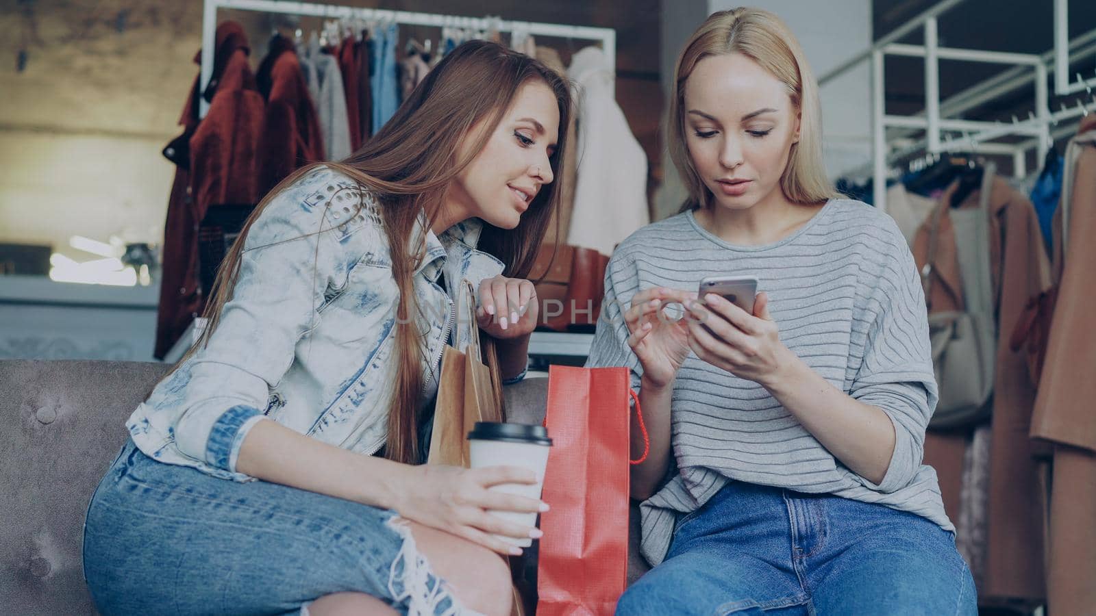 Cheerful young women chatting and using modern smartphone while sitting in nice clothing boutique. They are smiling and gesturing enthusiastically. by silverkblack