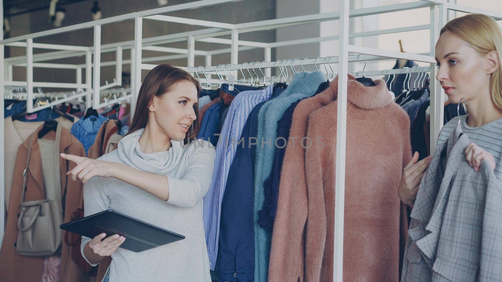 Young businesswoman is checking clothes in her clothing store with tablet in hands. Her employee is coming and asking about garment. Employer is talking to her and gesturing in friendly manner.