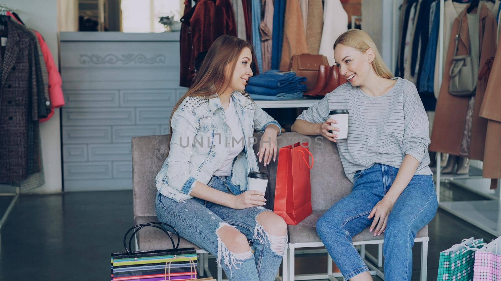 Two charming girls sitting in clothes shop with coffee, talking happily and laughing after shopping. Nice modern boutique with colourful garments, bags and shoes in background