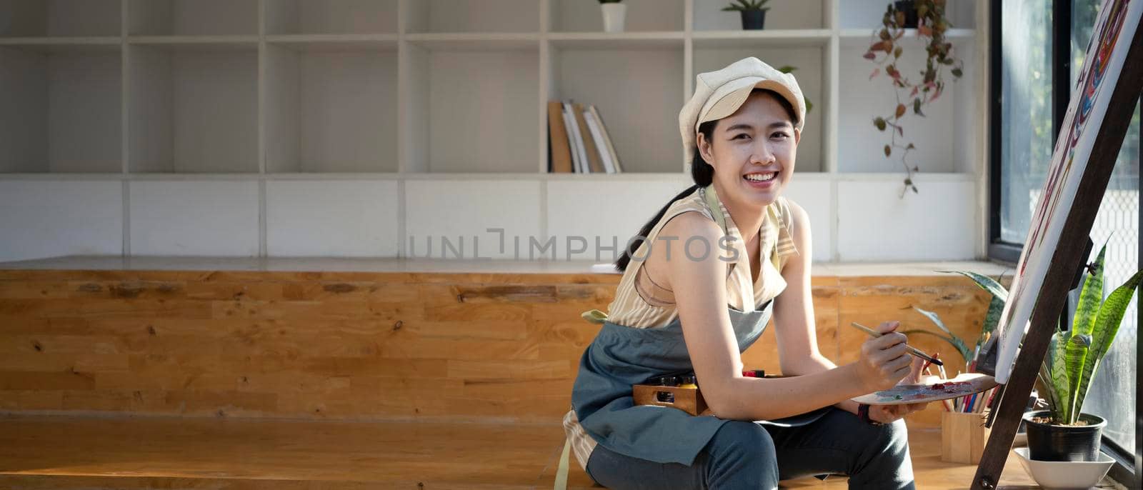 Beautiful female artist painting on canvas in art workshop and smiling to camera.