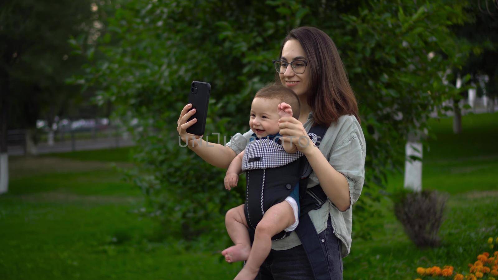 A young mother walks in the park with her little son and makes a video call on the phone. A girl in glasses walks with a newborn. A child in a kangaroo-backpack. 4k