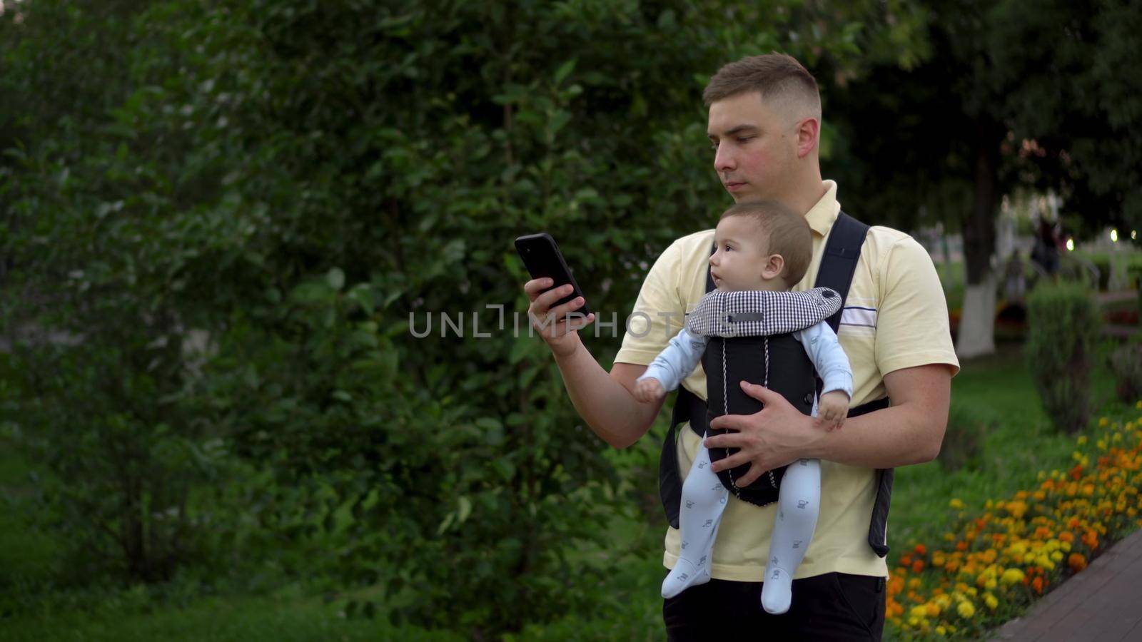 A young father walks in the park with a newborn child with a phone in his hands. The man is looking at the phone intently. Child in kangaroo-backpack. Closeup. by Puzankov