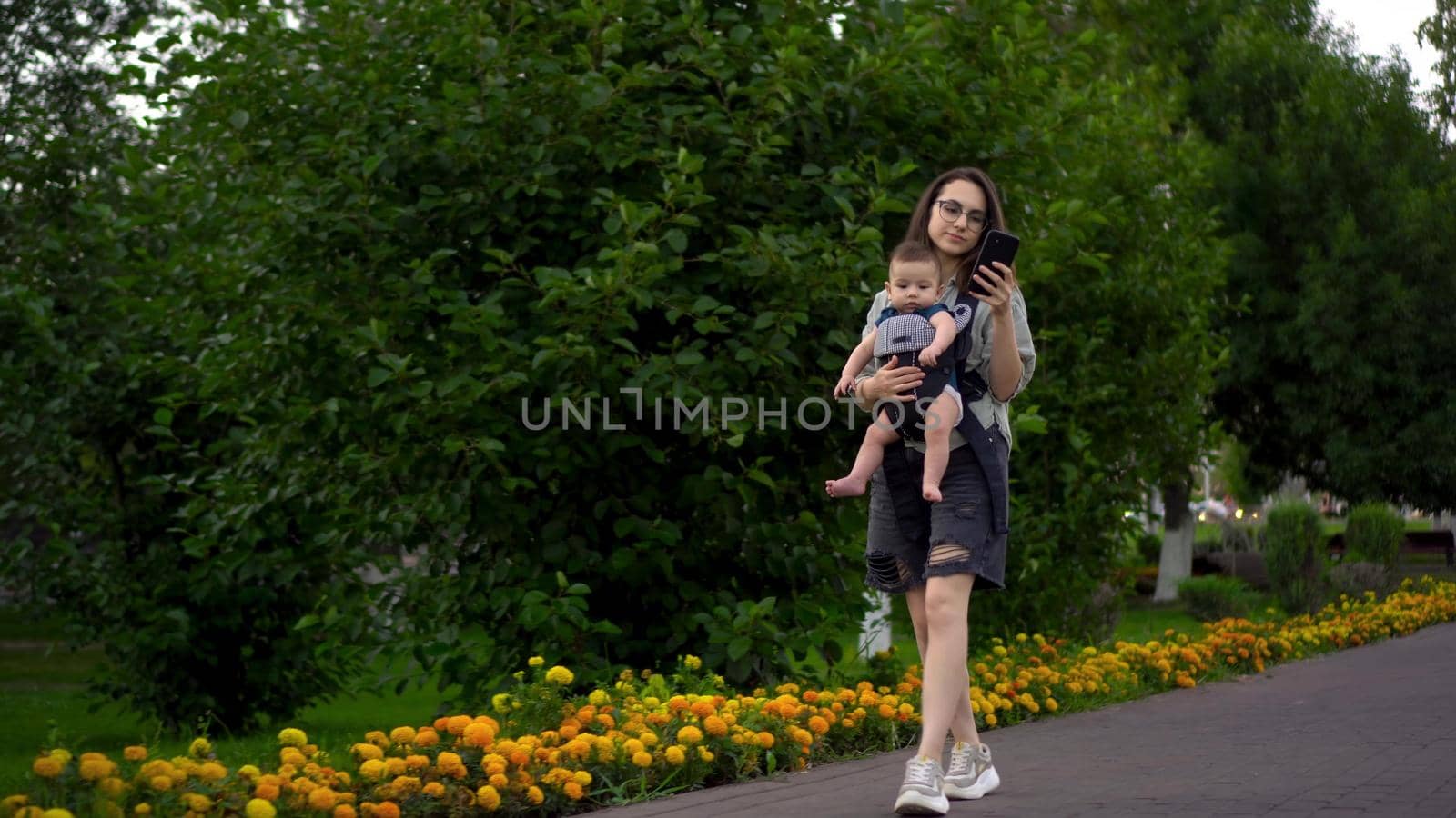 A young woman with her son walks along the alley in the park and texts on the phone. The girl in glasses walks. Newborn son in a kangaroo-backpack. 4k