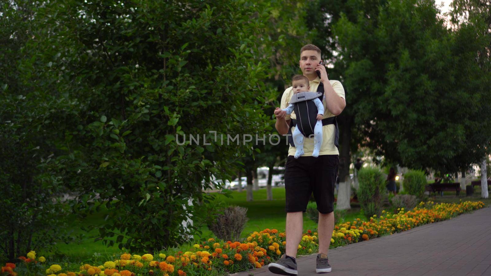 A young man walks in the park with a newborn child and speaks on the phone. The man is looking at the phone intently. A child in a kangaroo-backpack in his father's arms. by Puzankov