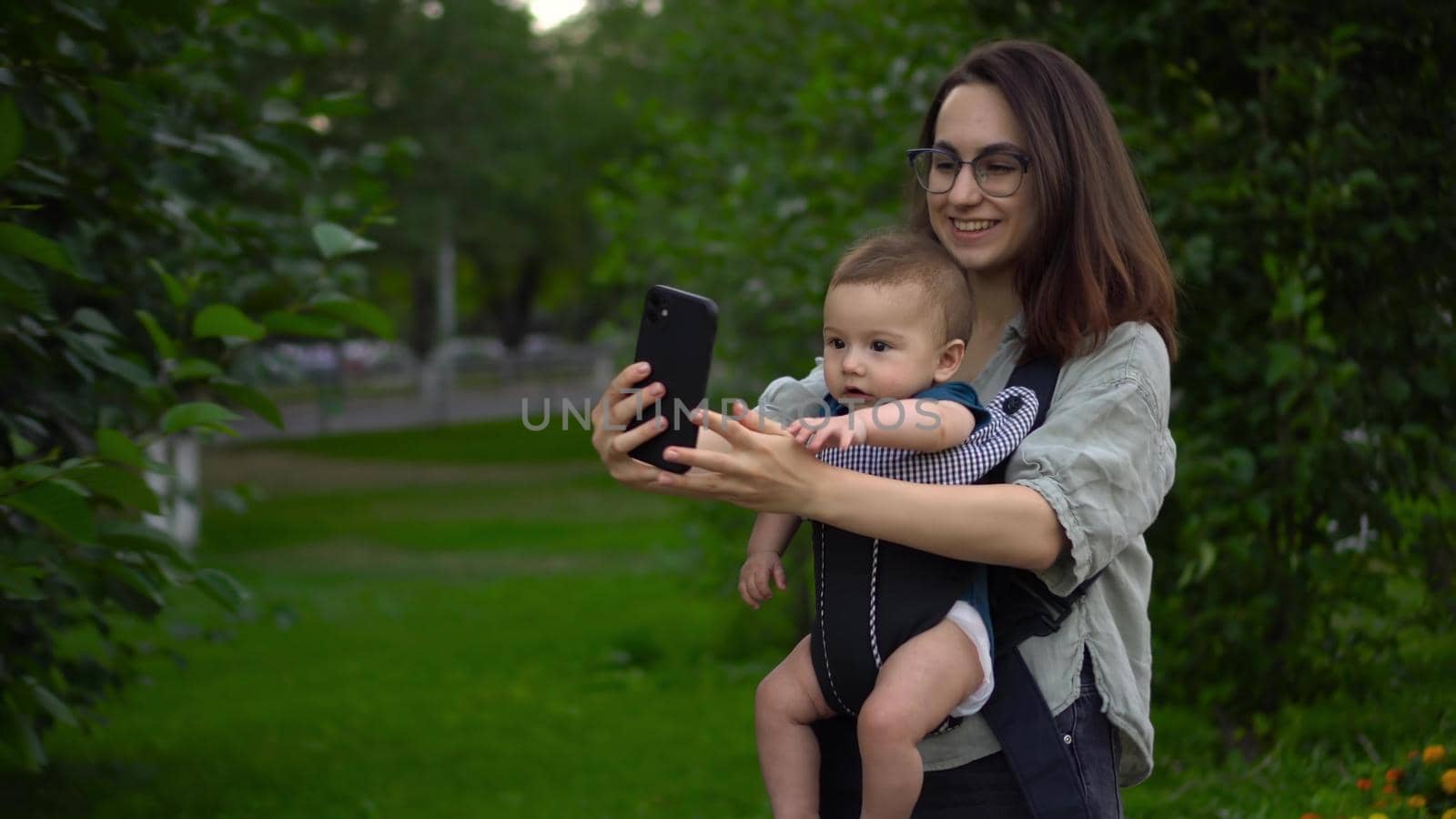 A young mother walks in the park with her little son and makes a video call on the phone. A girl in glasses walks with a newborn. A child in a kangaroo-backpack. 4k