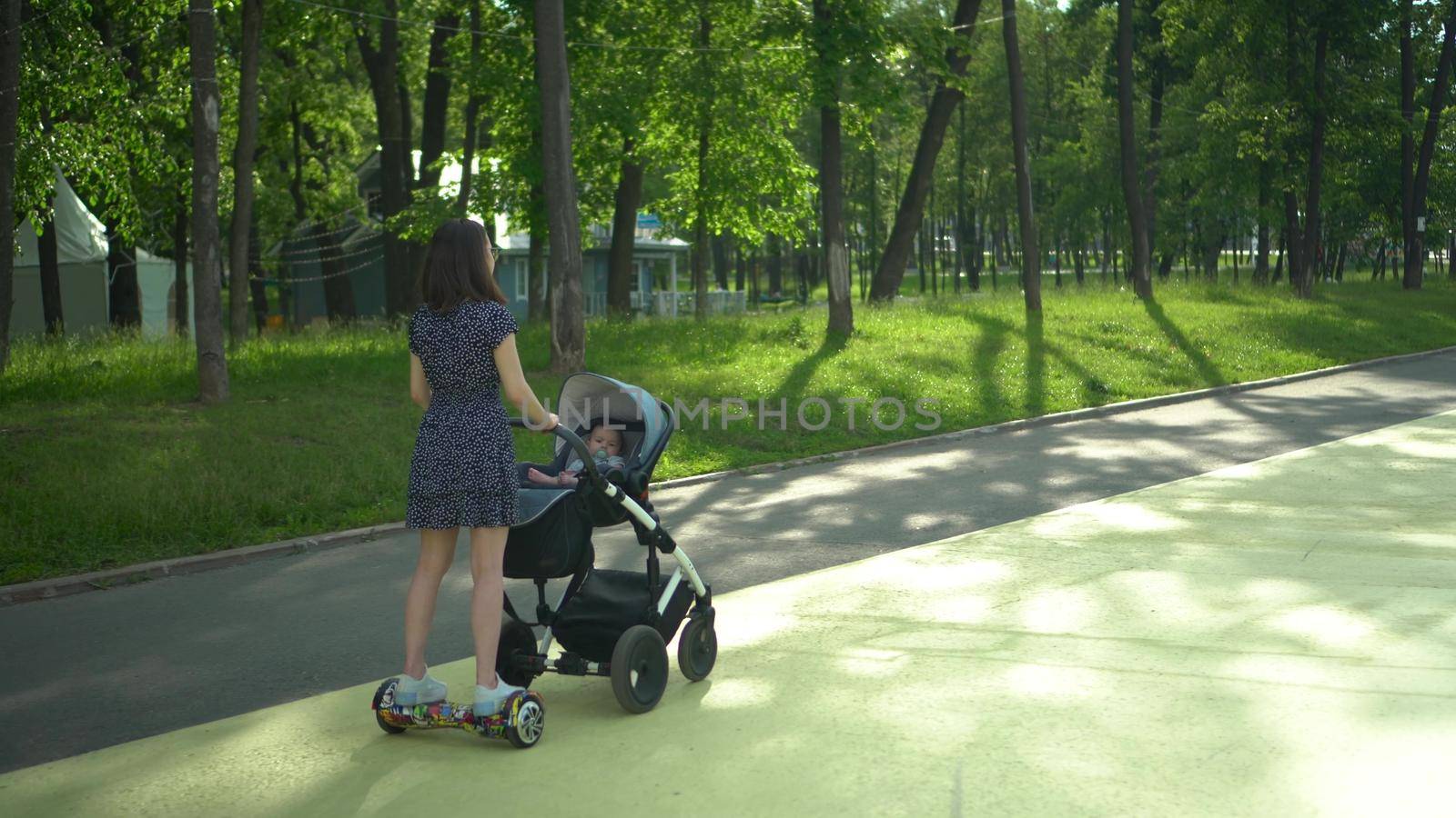 A young mother rides a gyro scooter and carries a stroller with a baby. Modern walk with a child in the park. Girl with glasses and a dress. 4k