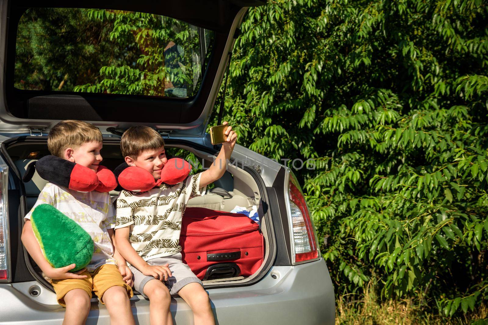 Two adorable little kids boy sitting in car trunk just before leaving for summer vacation. Sibling brothers making selfie on smartphone. Happy family going on long journey.