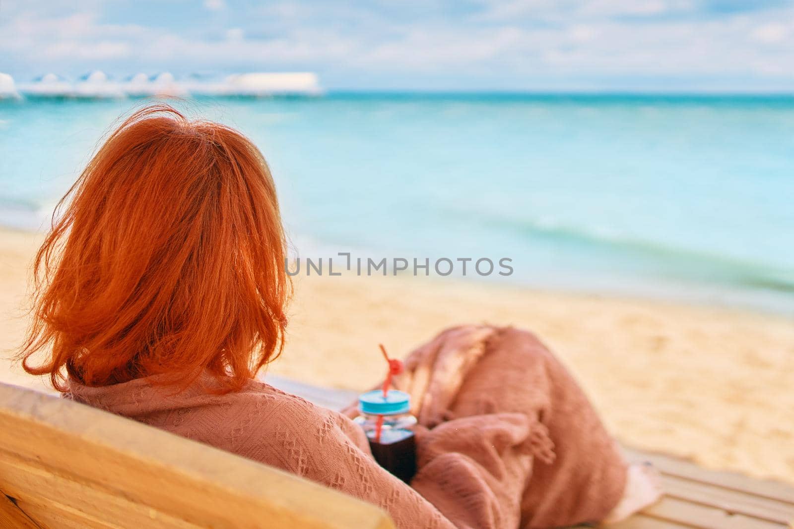Woman is resting on the sea beach and admiring the ocean landscape. Red-haired girl from behind is lying on chaise longue covered with a blanket. Alcoholic drink in female's hands. Summer holidays.