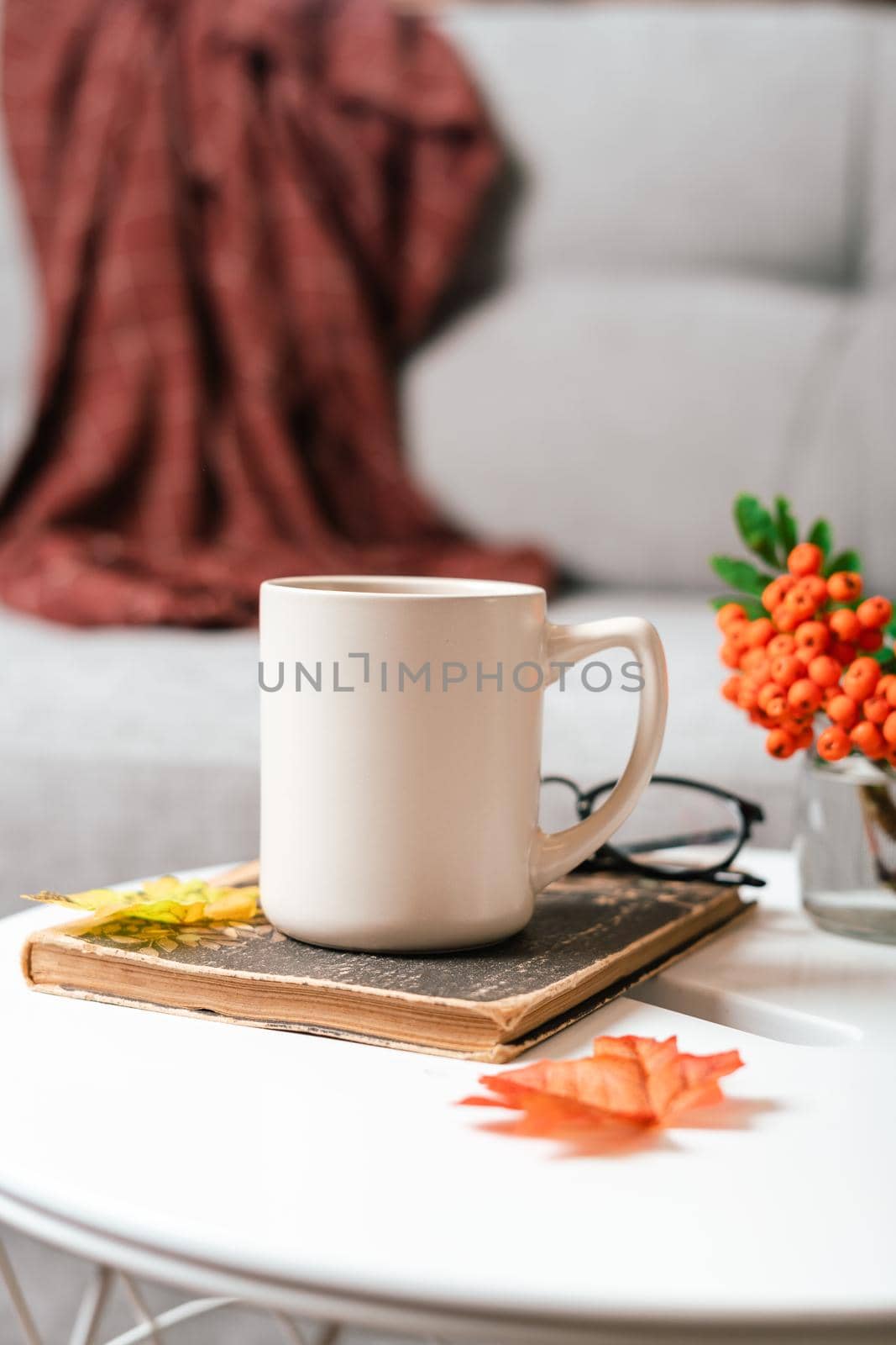 Still life book, candle, rowan berry and a cup of tea or coffee in the living room on a table, home decor in a cozy house. Autumn weekend concept, blanket and plaid. Fallen leaves and home decoration