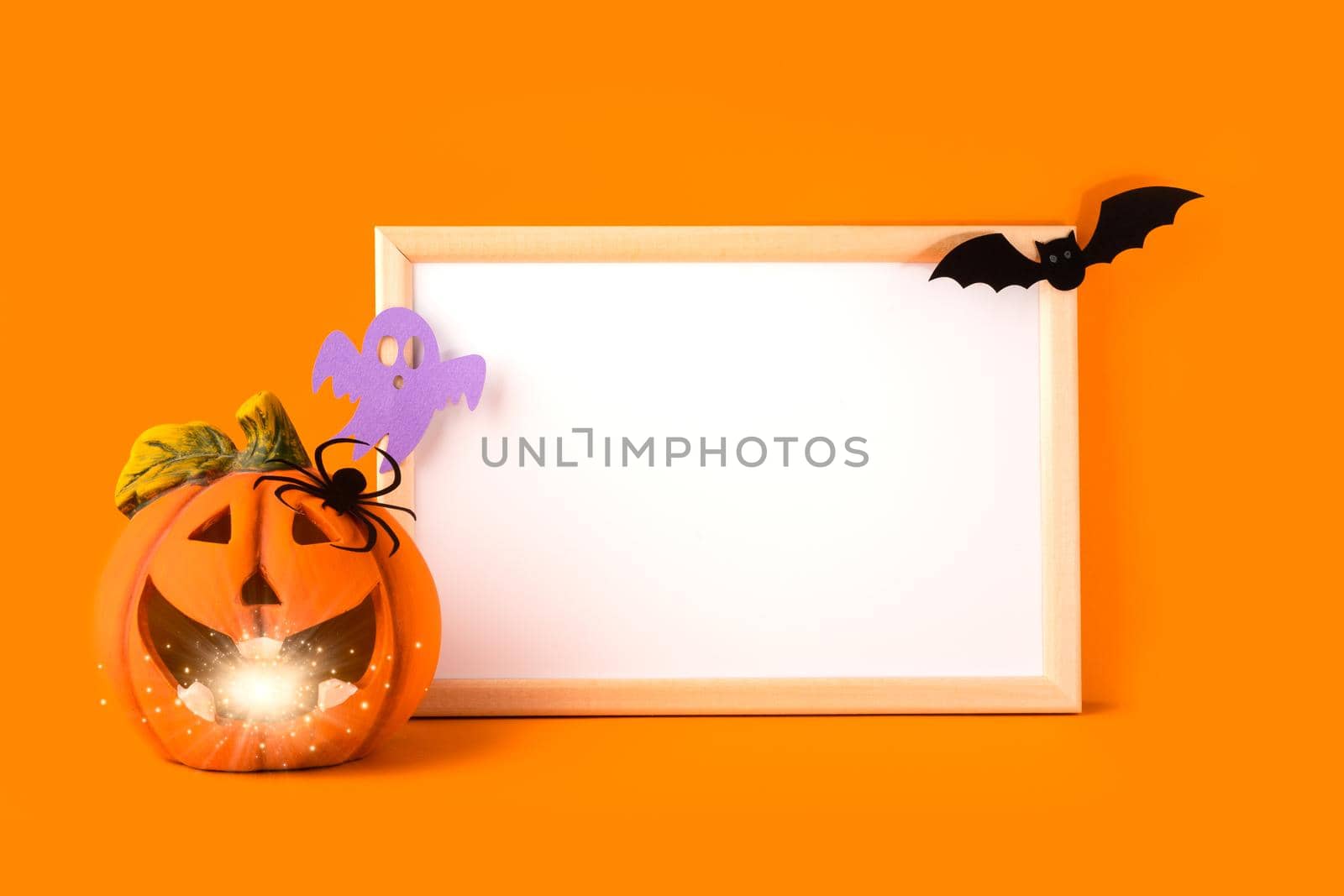 Halloween holiday concept. Jack o lantern, light, handmade paper decorations, spider, ghost, bat and blank frame on orange background. Halloween festival party, greeting card with mockup copy space.