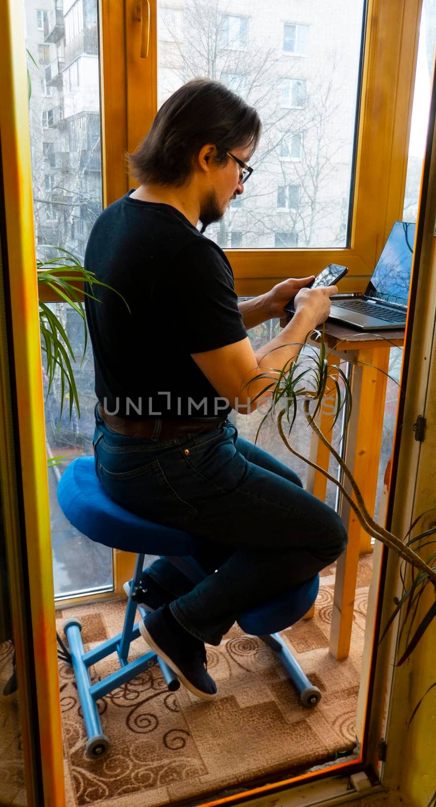 a man works at a laptop in a home office, sitting on a Orthopedic ergonomic kneeling chair on a glazed balcony among greenery. back health care