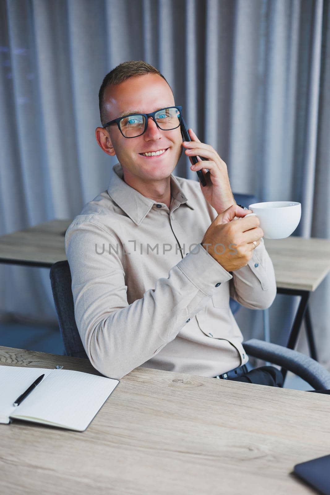 Indoor shot of cheerful bearded male freelancer work at distant, manages finances via smart phone, sits at wooden table, works with documentation, has happy expression. People, work, technology