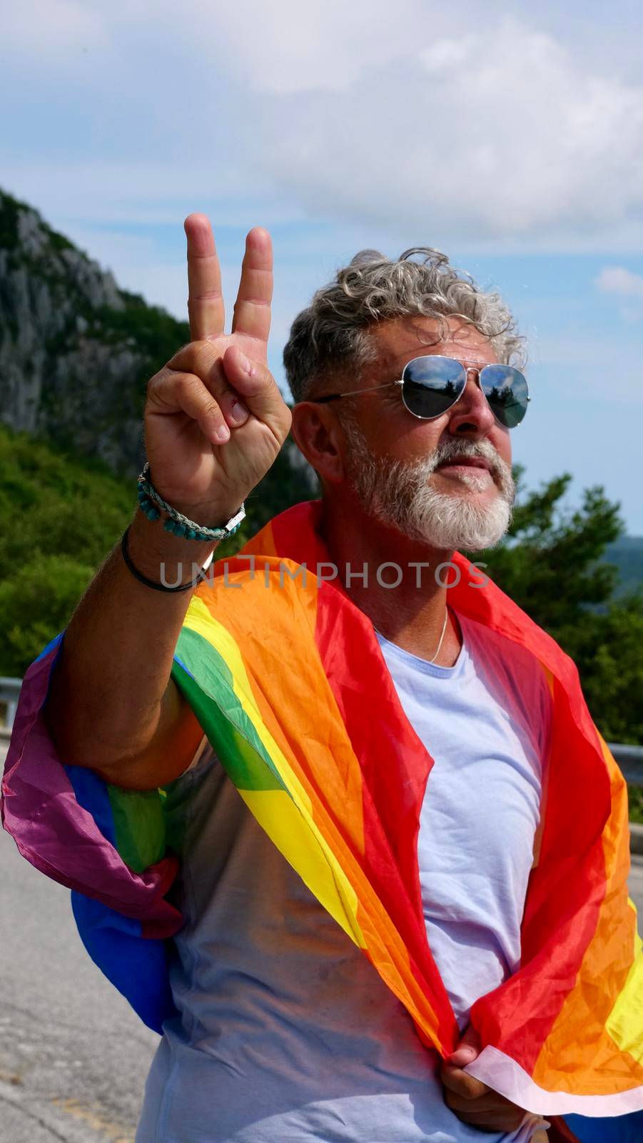 Portrait of a gray-haired senior elderly Caucasian man bisexuality with a beard and sunglasses with a rainbow LGBTQIA peace flag in mountains. Celebrates Pride Month, Rainbow Flag Day, gay parade