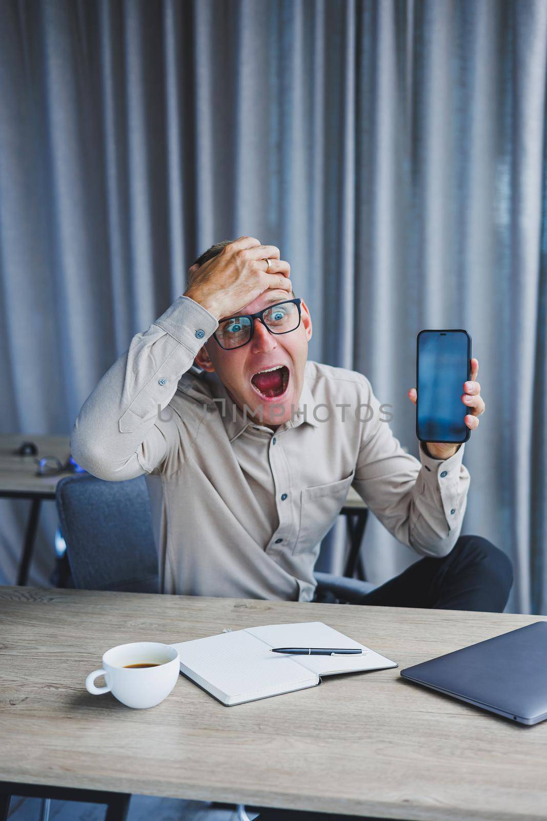 A young attractive male freelancer in glasses sits in the office and looks at the phone. Handsome blogger writing ideas on phone while sitting at wooden table in office with laptop
