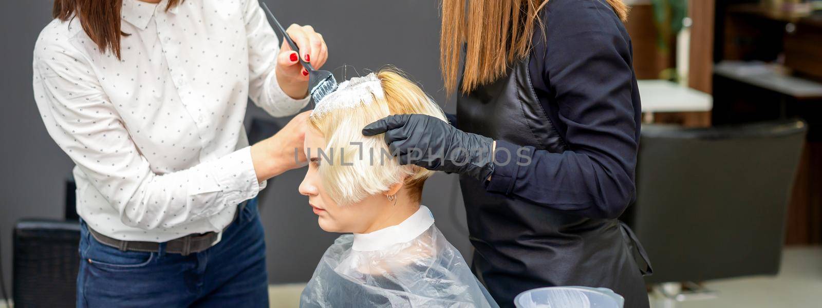 Two female hairdressers dyeing hair of young caucasian woman in hair salon
