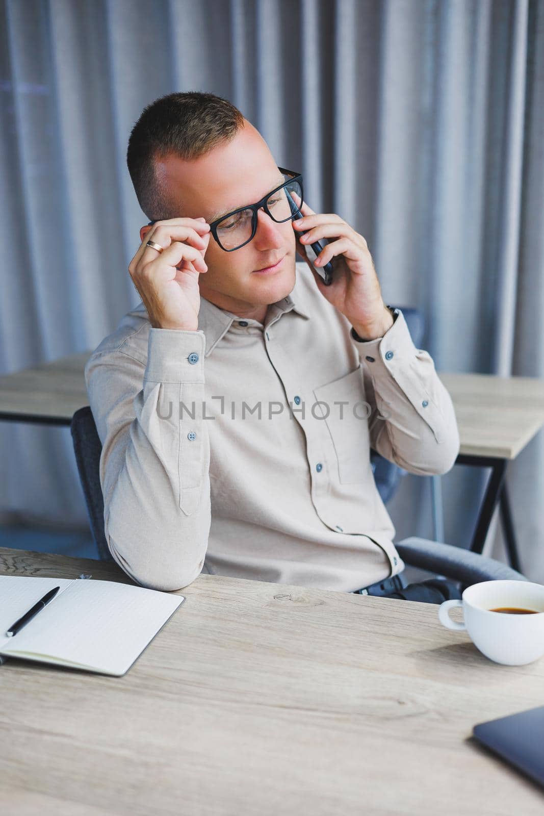 Indoor shot of cheerful bearded male freelancer work at distant, manages finances via smart phone, sits at wooden table, works with documentation, has happy expression. People, work, technology