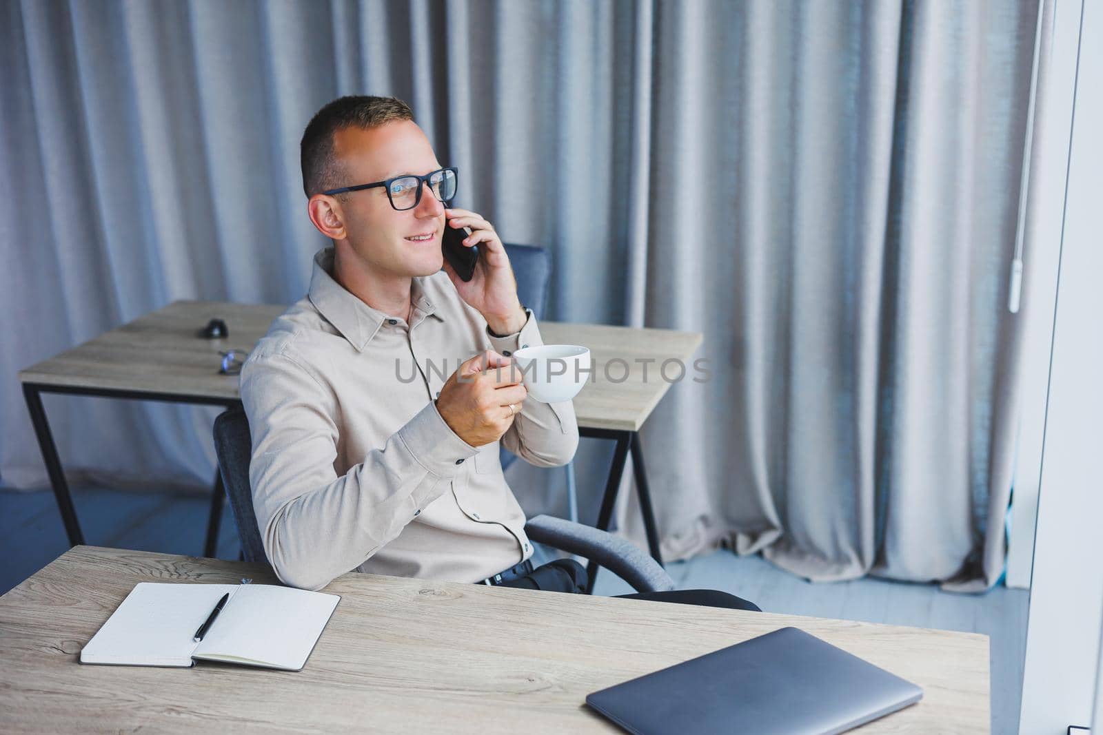 Indoor shot of cheerful bearded male freelancer work at distant, manages finances via smart phone, sits at wooden table, works with documentation, has happy expression. People, work, technology
