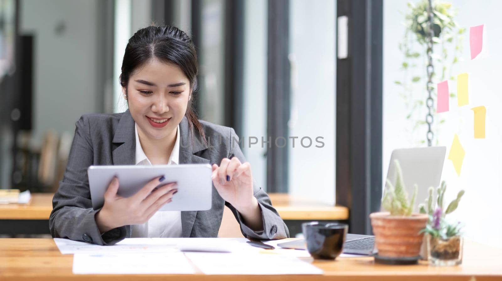 Beautiful smiling Asian businesswoman wearing glasses hand holding pen working using tablet at office. by wichayada