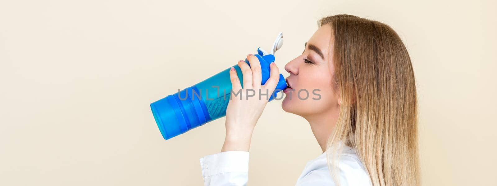 Side view of beautiful young caucasian woman is drinking water from plastic bottle against a beige background