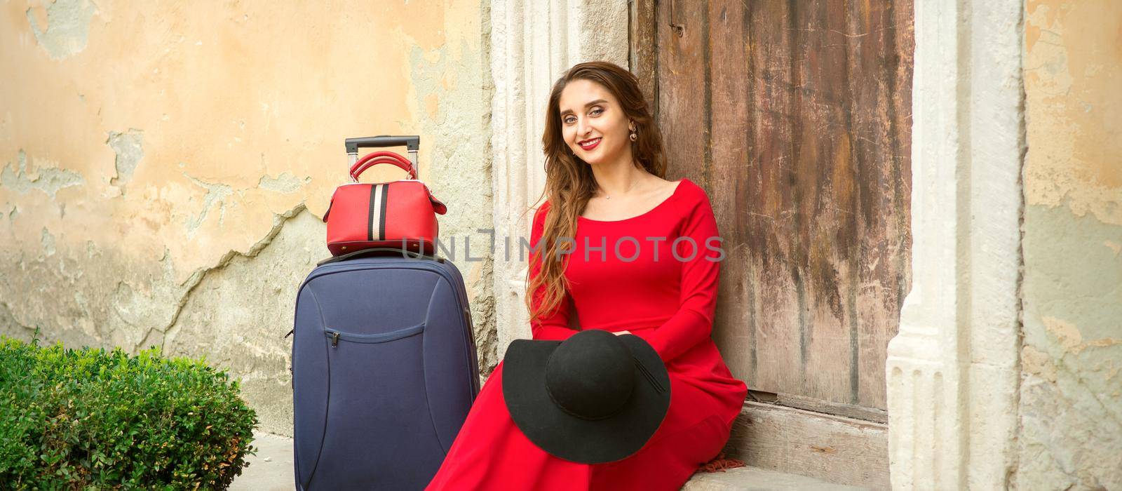 A young caucasian woman is sitting on the threshold of an old house in a red long dress with suitcase looking at the camera outdoors