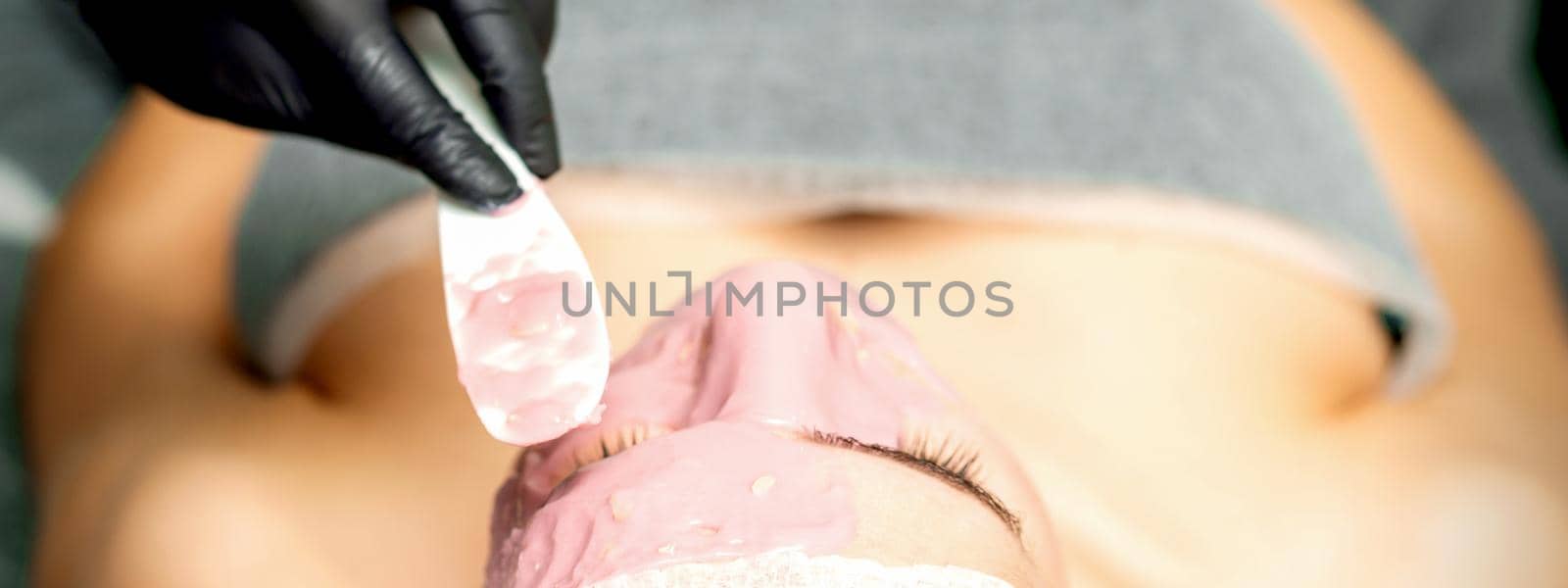 The cosmetologist applying an alginic mask to the face of a young woman in a beauty salon