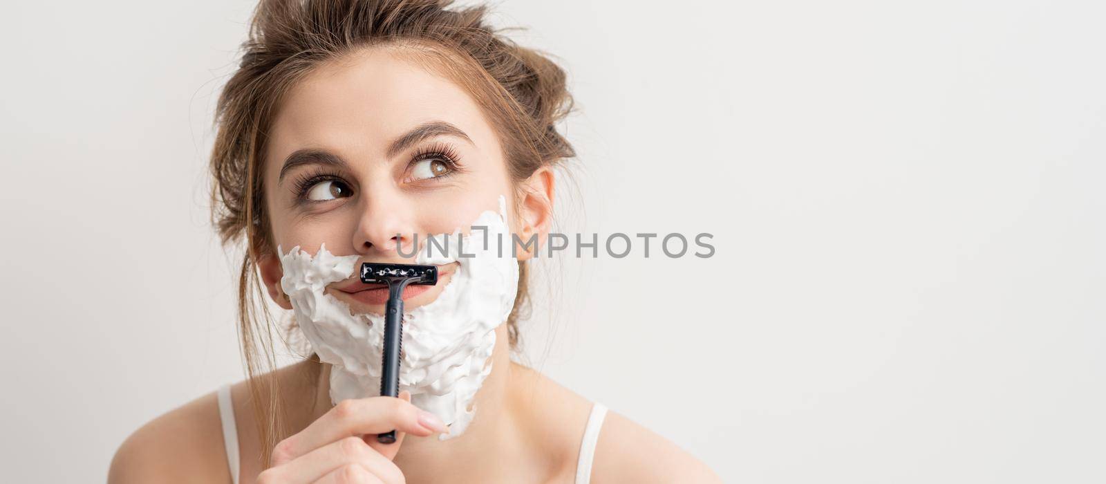 Beautiful young caucasian smiling woman shaving her face with razor looking up on white background