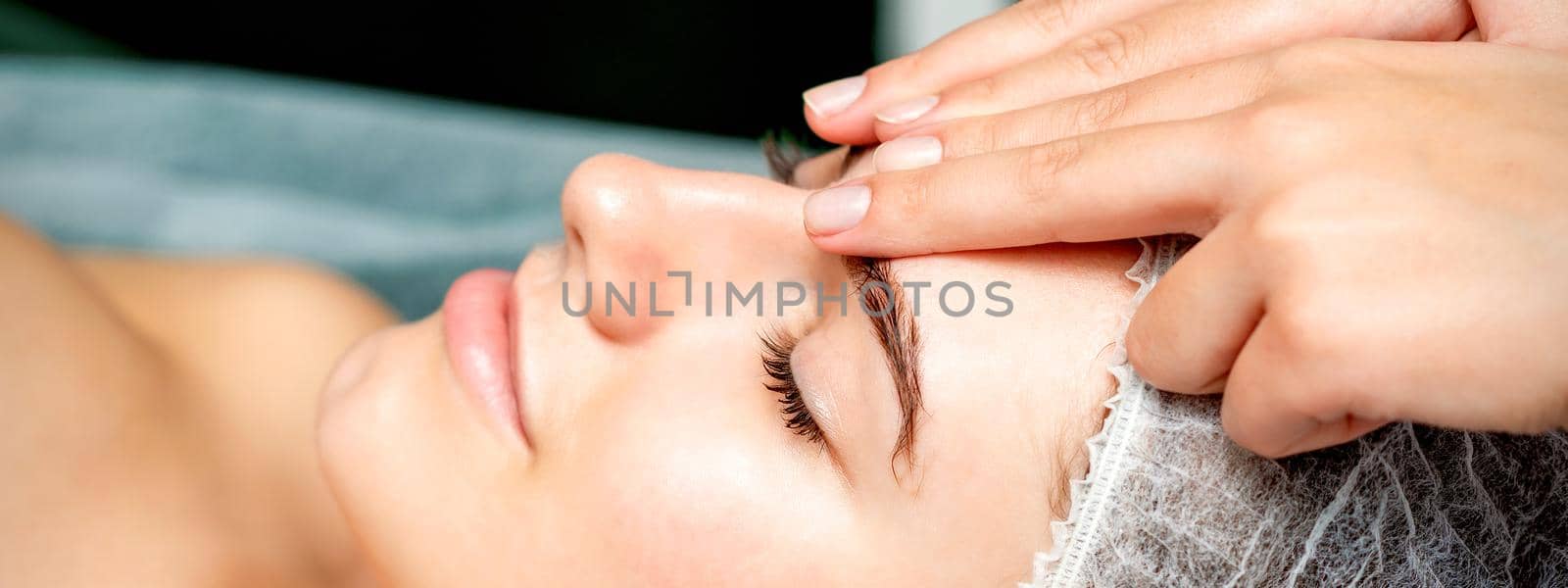 Side view fingers of female therapist making anti-age massage of forehead wrinkles to young caucasian woman in spa beauty salon