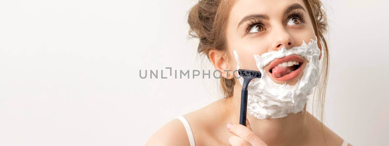 Beautiful young caucasian smiling woman shaving her face with razor sticking out tongue looking up on white background