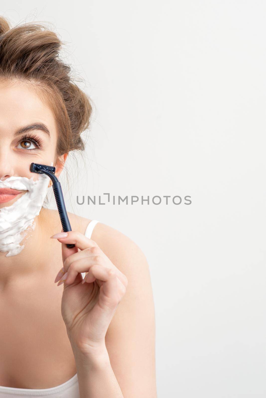 Beautiful young caucasian smiling woman shaving her face with razor looking up on white background