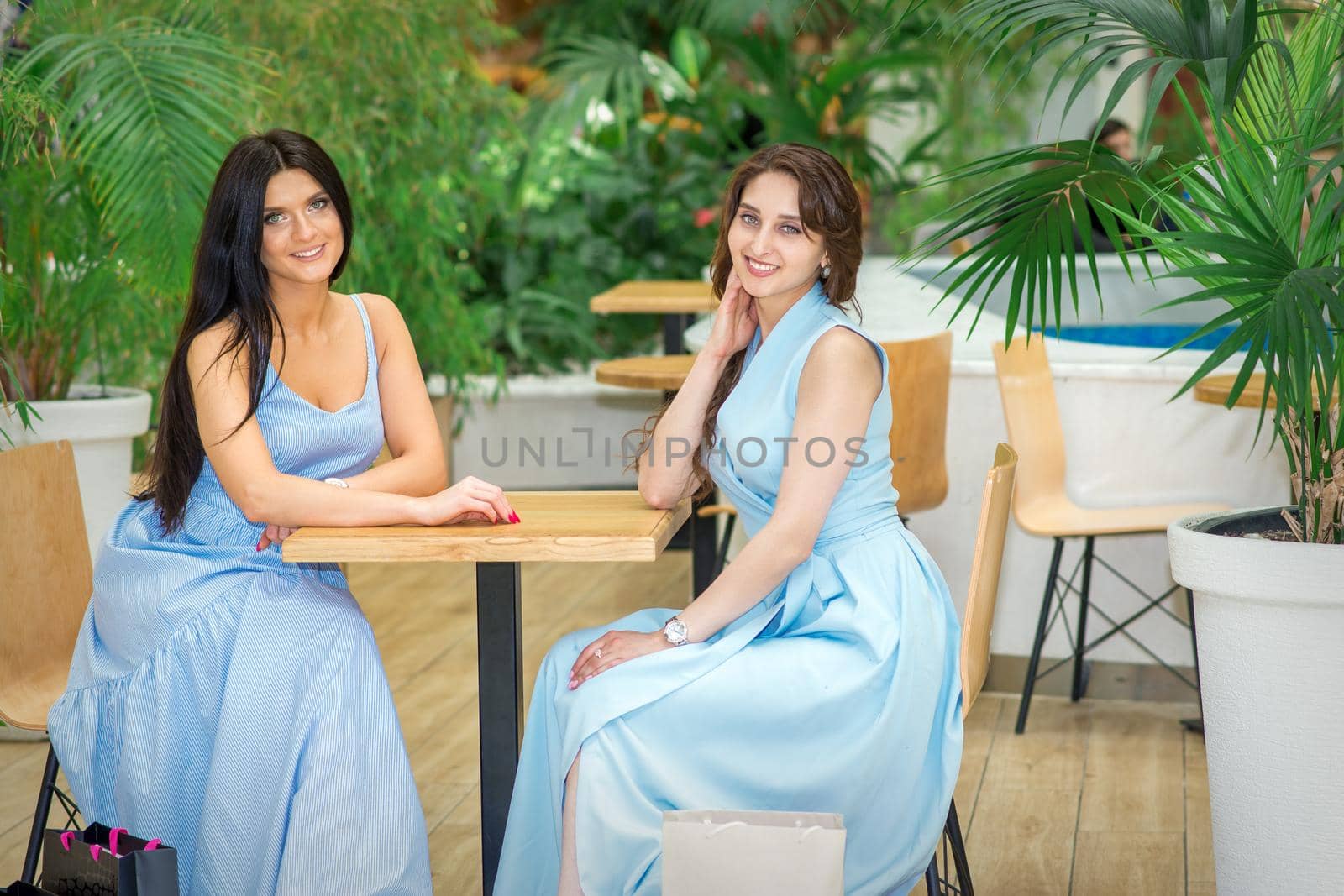 Two beautiful smiling caucasian young women in long blue dresses sitting at the table and looking at the camera in a cafe outdoors