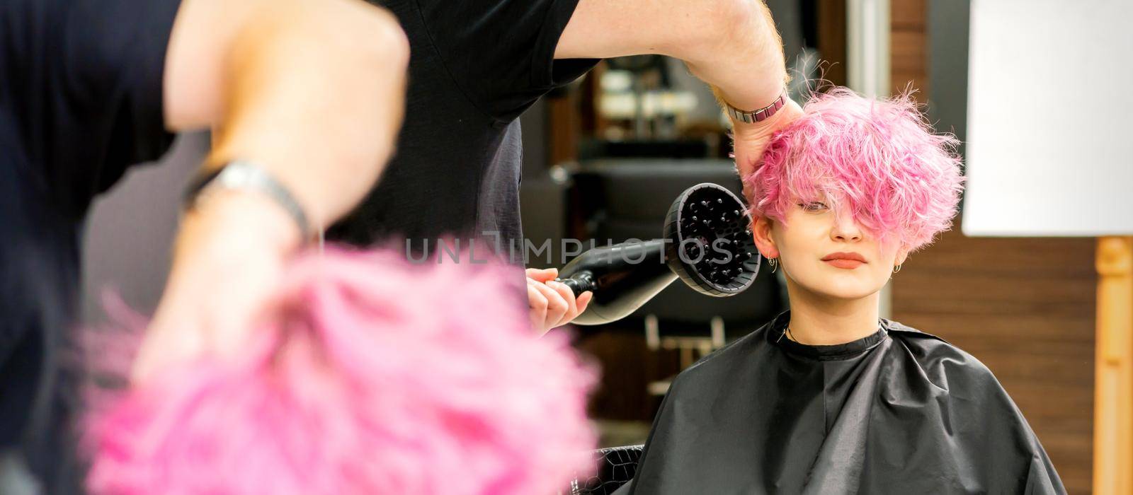 A male hairdresser professional drying stylish pink hair of the female client with a blow dryer in a beauty salon
