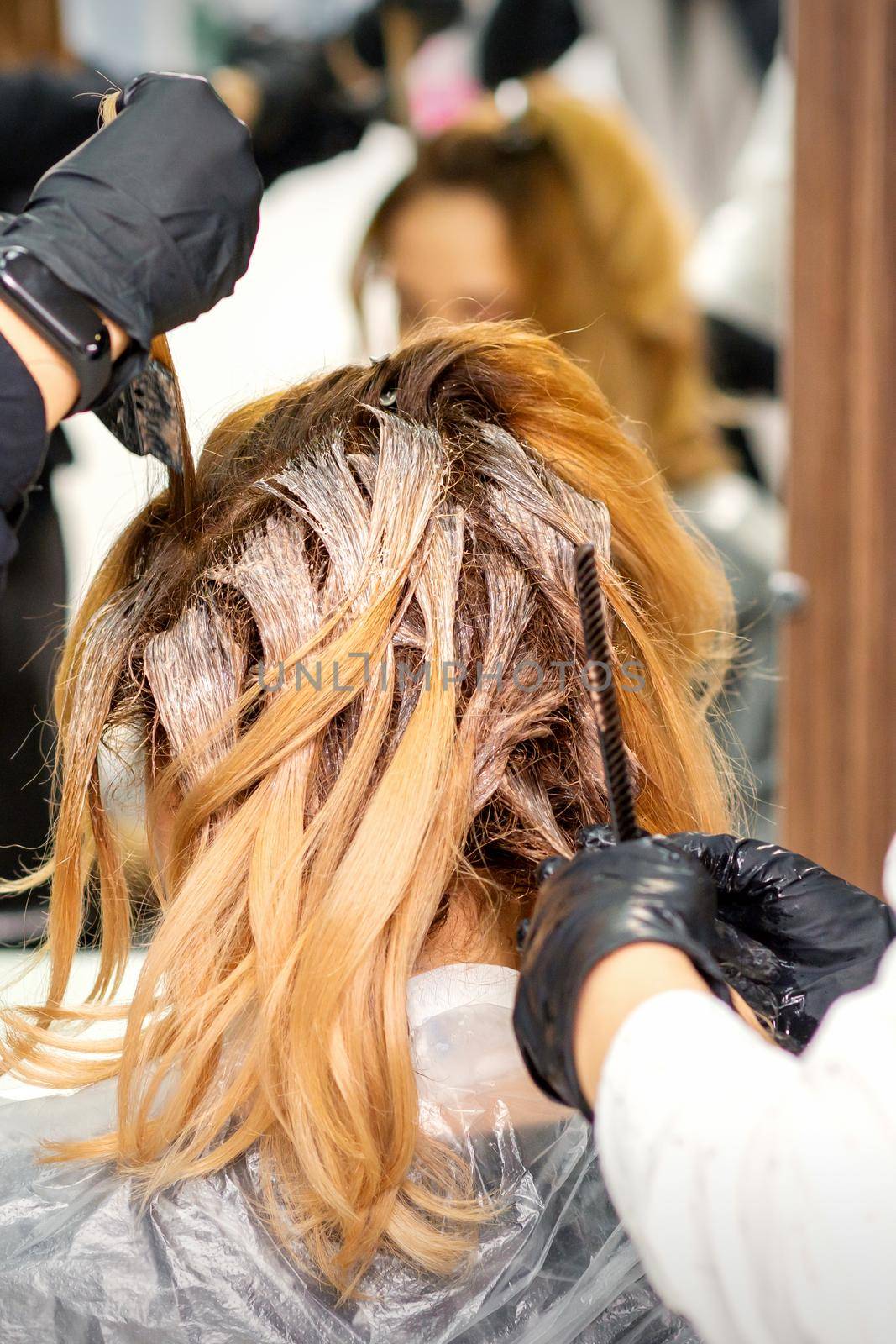 Back view of a female hairdresser in gloves coloring hair of redhead young woman with light or white dye in a beauty salon