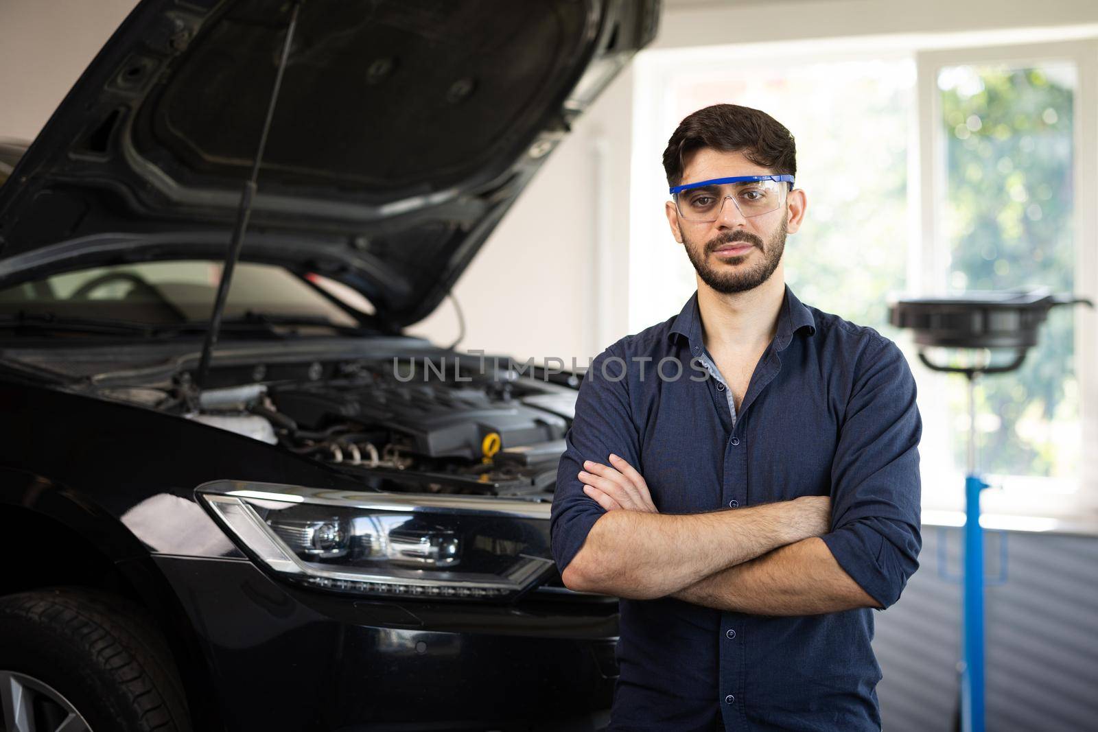 Portrait Bearded Handsome Car Mechanic is Posing in a Car Service. He Wears a Jeans Shirt and Safety Glasses. His Arms are Crossed. Specialist Manager Looks at a Camera and Smiles by uflypro