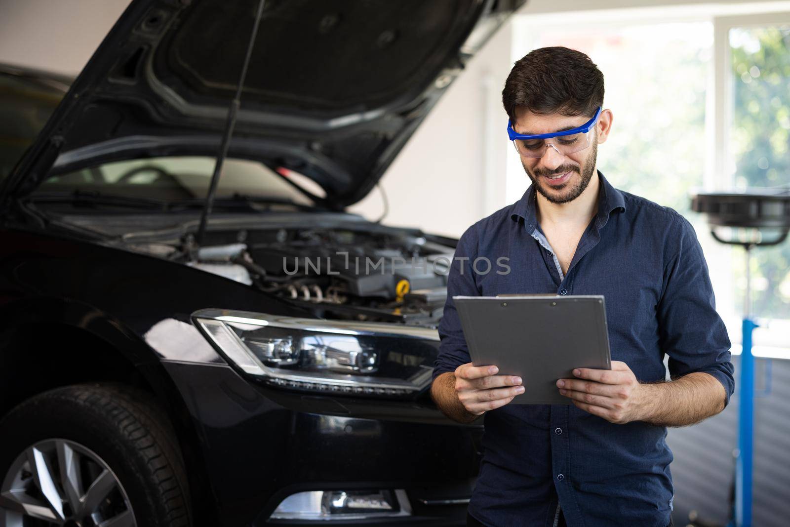 Mechanic inspects the car undercarriage way and makes a note on his inspection sheet. Automobile service, car mechanic. Car service employe inspect car. Modern workshop. by uflypro