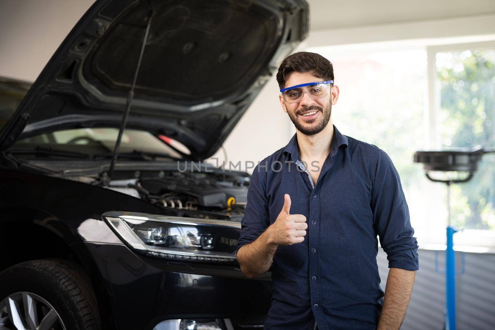 Happy technician bearded brunet mechanic man 20s in safety glasses work in vehicle repair shop show thumb up like gesture on light workshop background indoors by uflypro