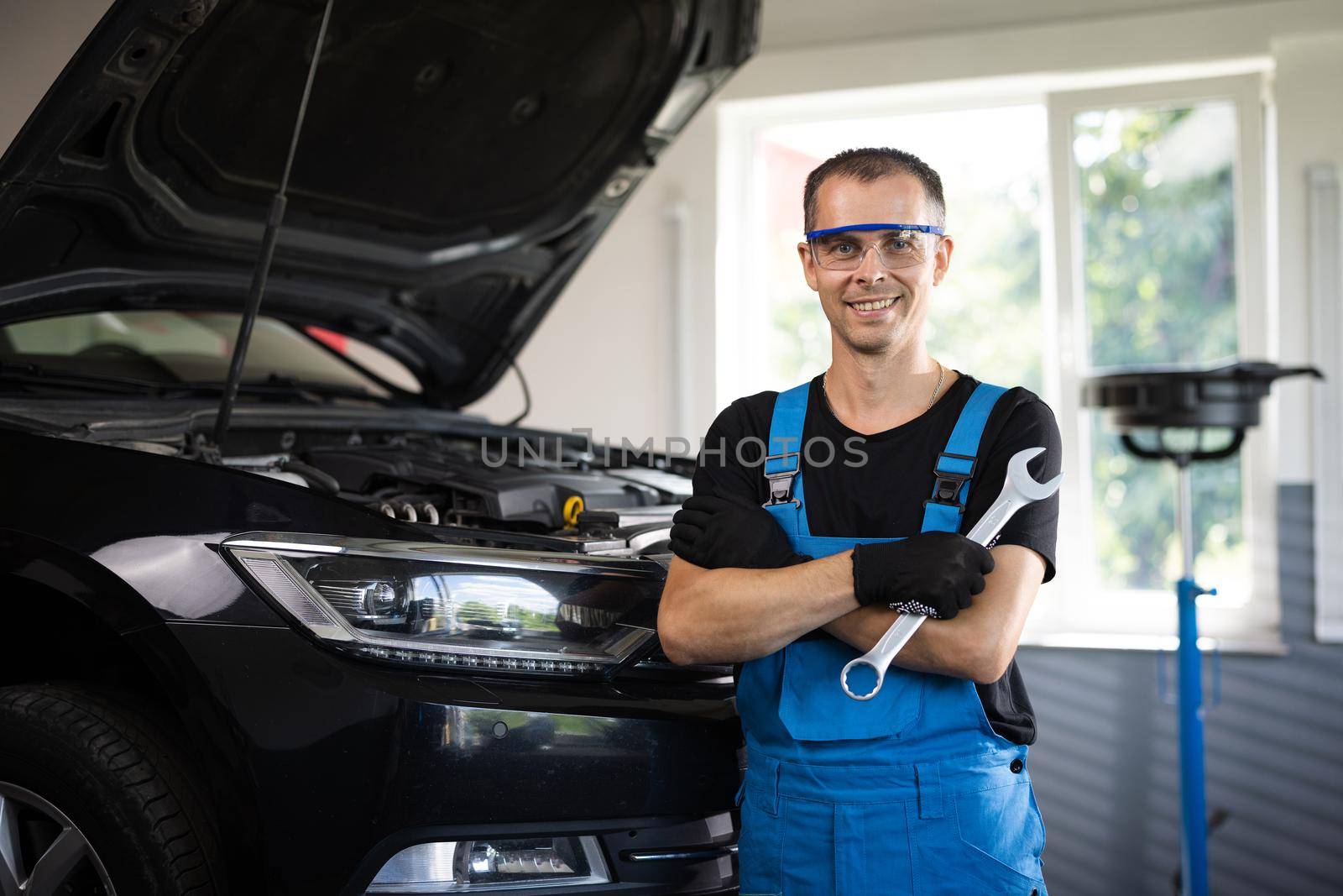 Portrait of handsome automotive mechanic man in blue uniform standing in garage. Vehicle service manager worker work in mechanics workshop, looking at camera after check the engine.