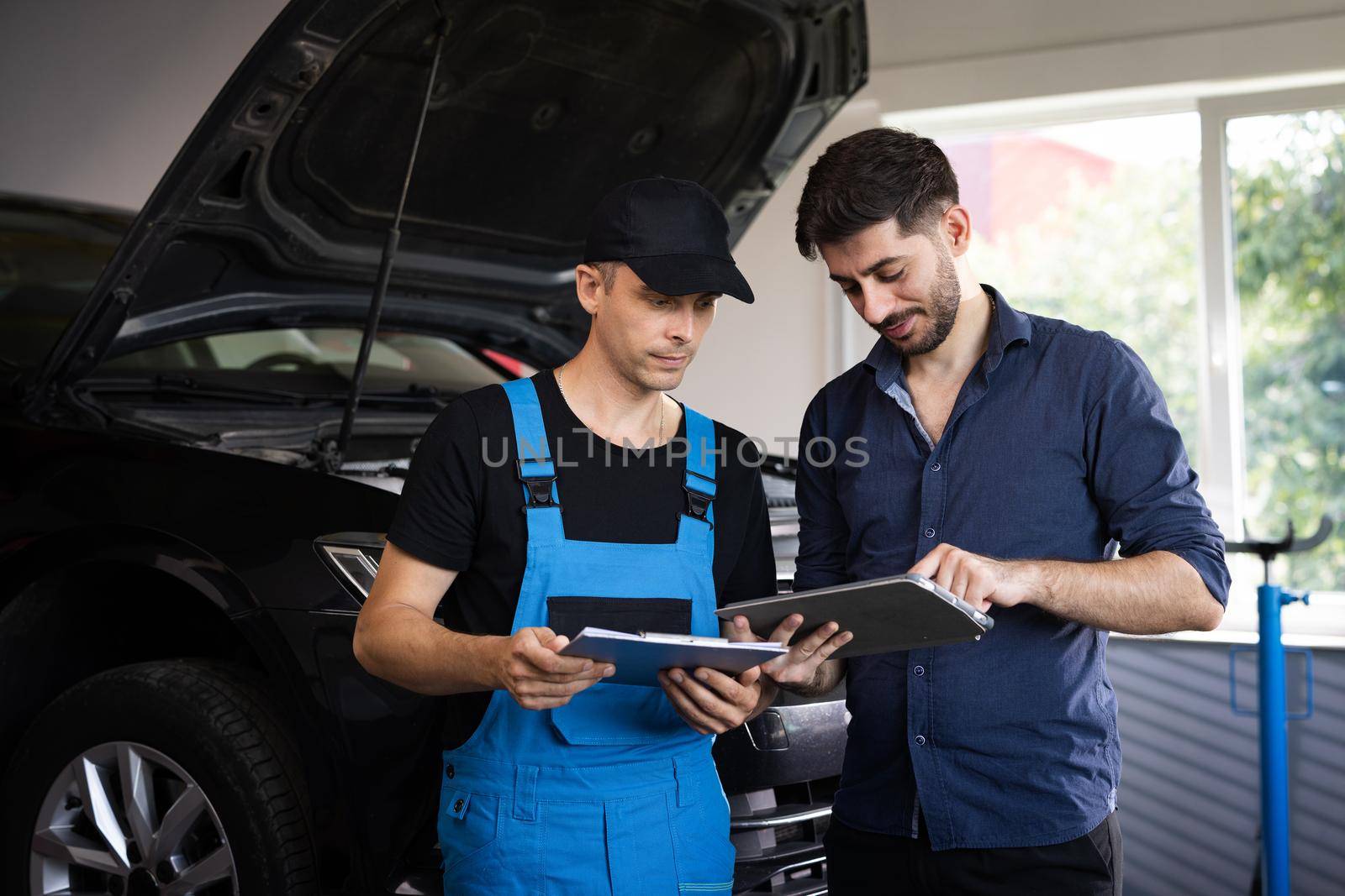 Customer and mechanic discuss upcoming car repair. Auto service, repair, deal and people concept. Mechanic and car owner signing paper at workshop. by uflypro