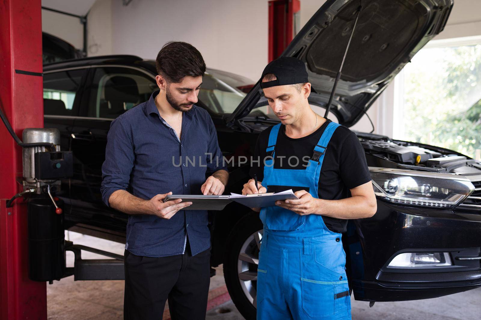 Manager Checks Data on a Tablet Computer and Explains the Breakdown to Mechanic. Car Service Employees Inspect the Bottom and Skid Plates of the Car. Specialist is Showing Info on Tablet. by uflypro