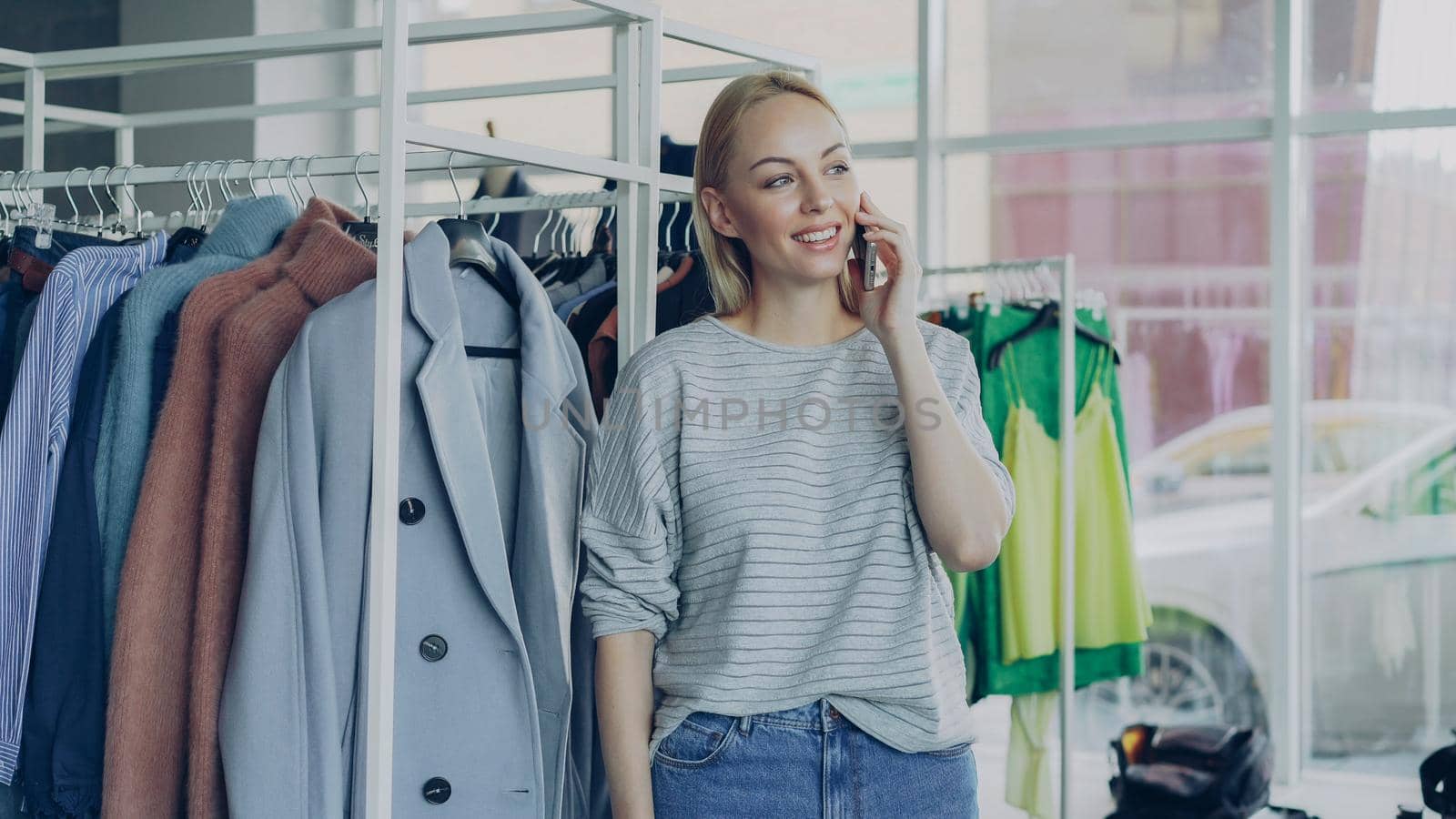 Cute blond girl is chatting on smart phone in boutique, touching clothes on hangers and smiling. Lots of fashionable clothes and large window in background. by silverkblack