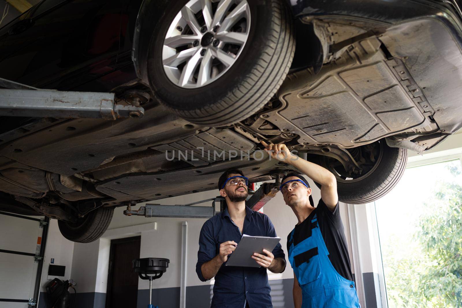 Manager Checks Data on a Notebook and Explains the Breakdown to a Mechanic. Car Service Employees Inspect the Bottom and Skid Plates of the Car. Modern Workshop. Auto Service by uflypro