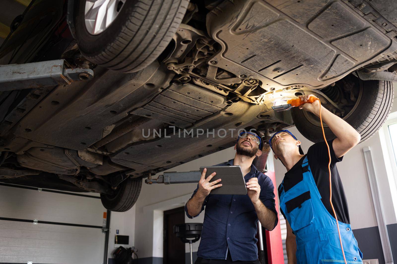 Manager Checks Data on a Tablet Computer and Explains the Breakdown to Mechanic. Car Service Employees Inspect the Bottom of the Car with a LED Lamp. Modern Workshop. Auto Service.