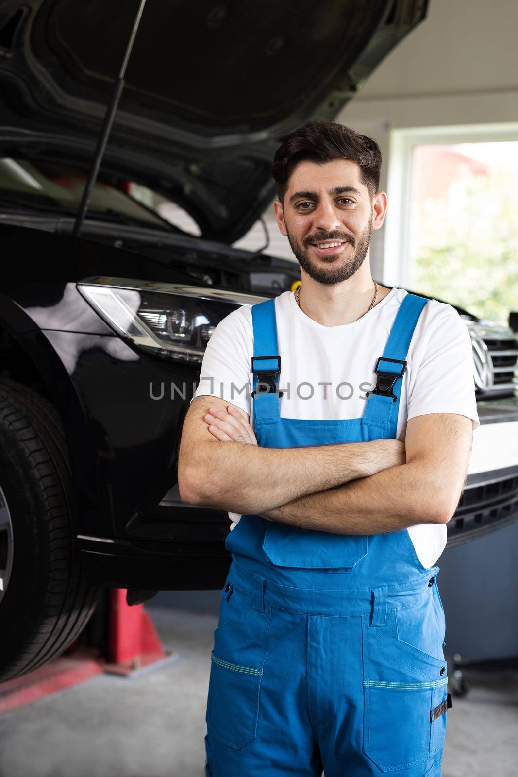 Male car mechanic at workplace in spacious repair shop. Portrait of bearded car mechanic crosses hands in a car workshop in blue uniform with equipment looking into camera.