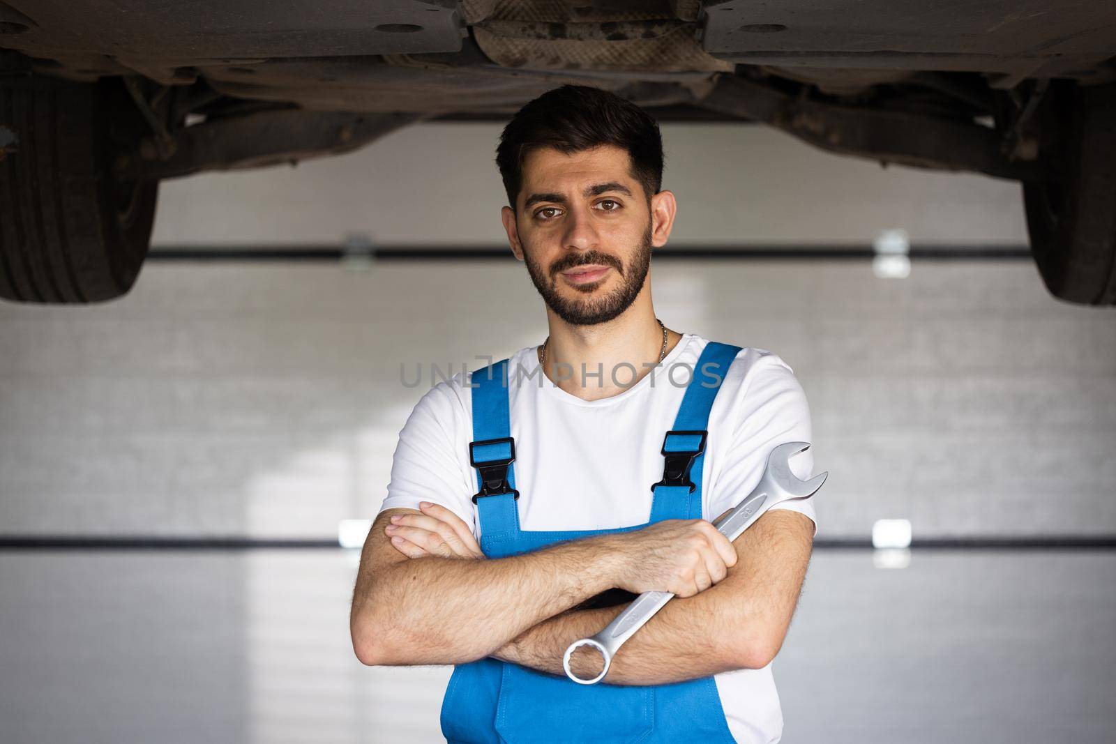 Portrait of bearded handsome automotive mechanic man standing in garage. Vehicle service manager worker work in mechanics workshop looking at camera after check and maintenance to repair the engine by uflypro