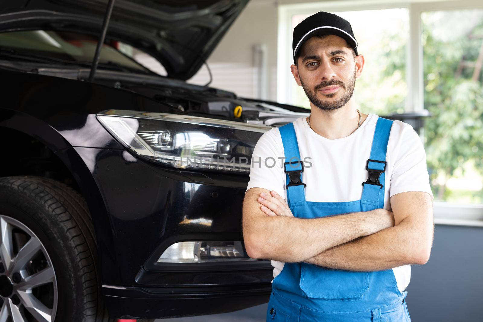 Young caucasian bearded man in blue overalls and black cap looks into camera, while smiling and crosses arms. Male car mechanic at workplace in spacious repair shop.