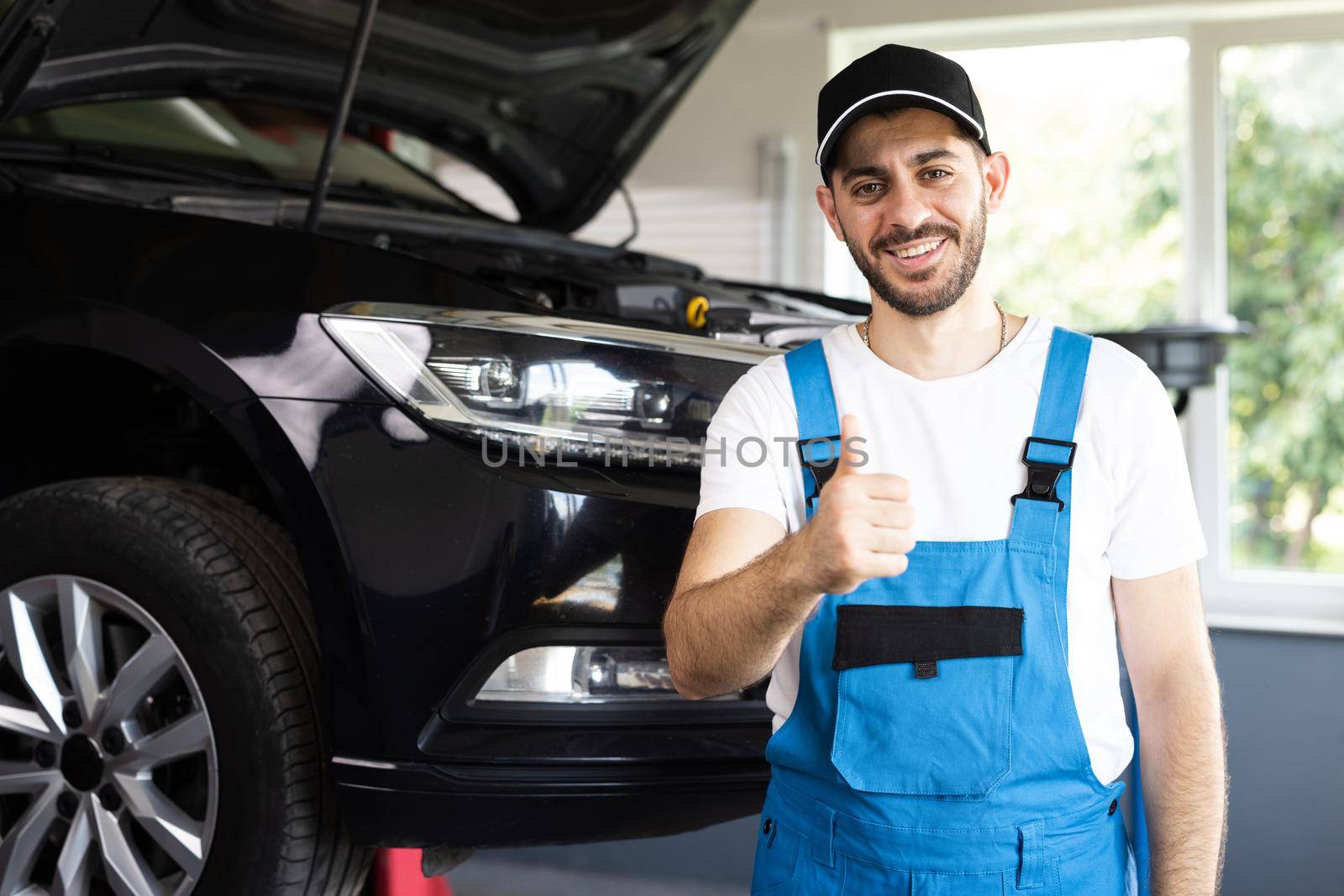 Portrait of bearded car mechanic in a car workshop shows thumbs up. Positive auto service worker in blue overalls and cap smiling to camera and showing thumb up gesture, approving car repair workshop by uflypro