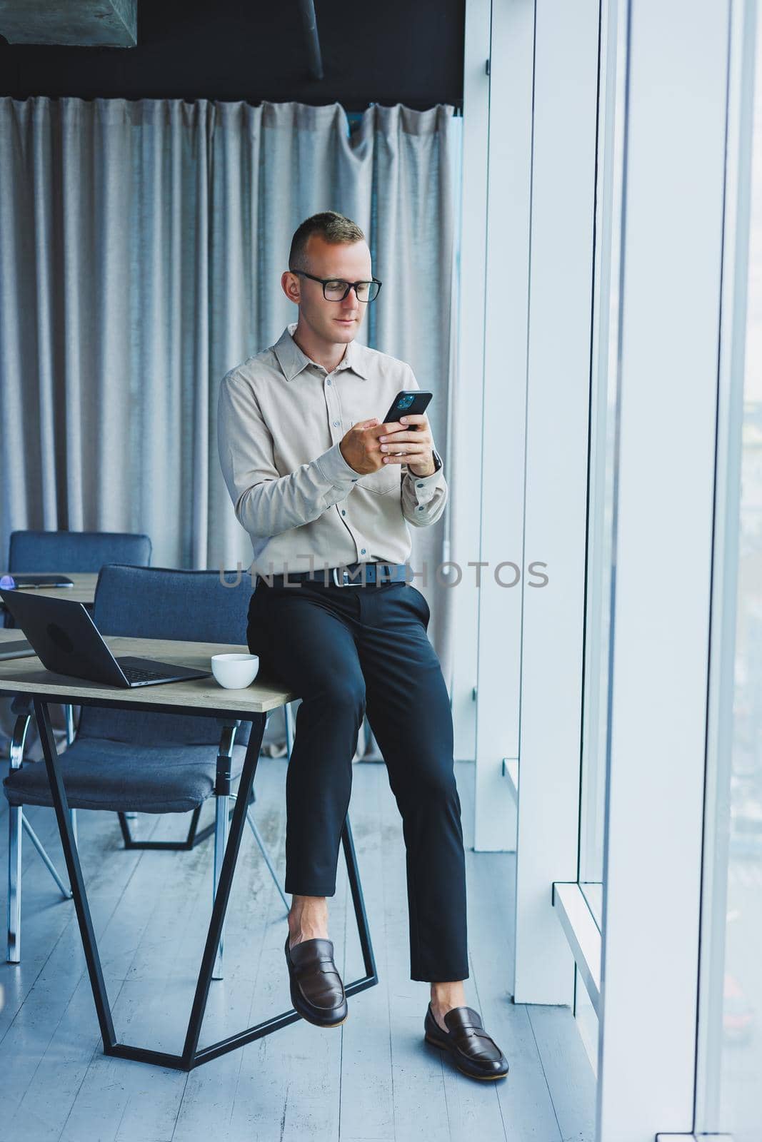 Young smiling businessman holding a cup of coffee in his hands in the office businessman looking out the window standing in a modern workplace man taking a vacation from hard work